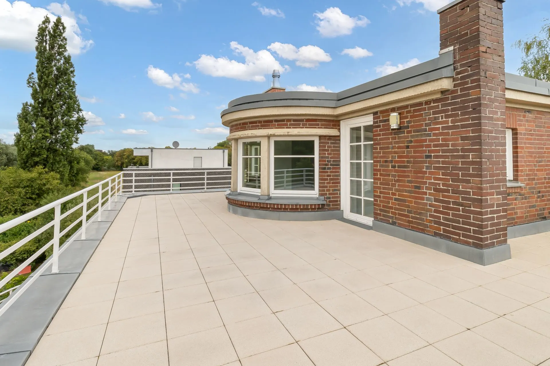 Große Dachterrasse mit Geländer an einem Backsteinhaus, Fenster und Tür zur Terrasse sichtbar.
