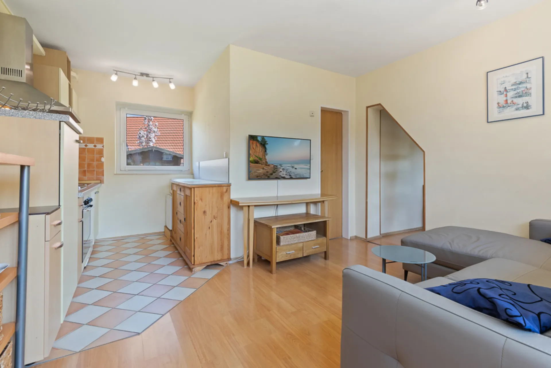 Wohnzimmer mit grauem Sofa, Fernseher, Holztisch und offener Küche mit Fliesenboden.
