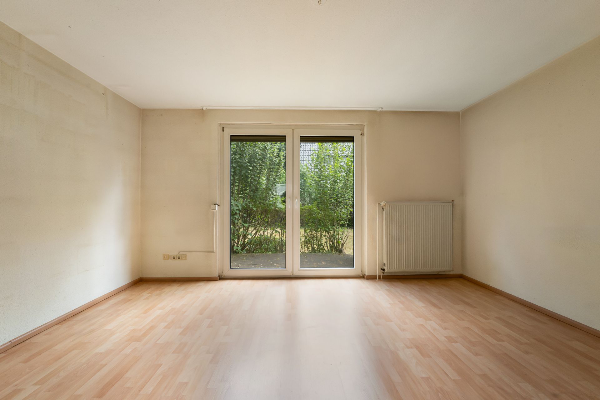 Leeres Zimmer mit heller Holzboden, Fenstertür zum Garten und Heizkörper an der rechten Wand.