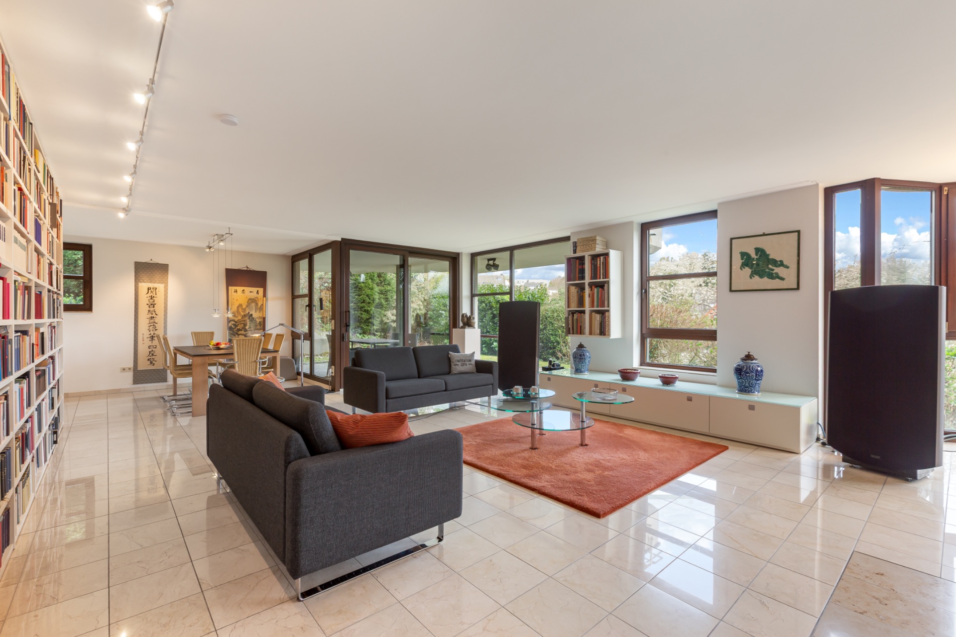 Helles Wohnzimmer mit zwei Sofas, Glastisch, Bücherregal und großen Fenstern.