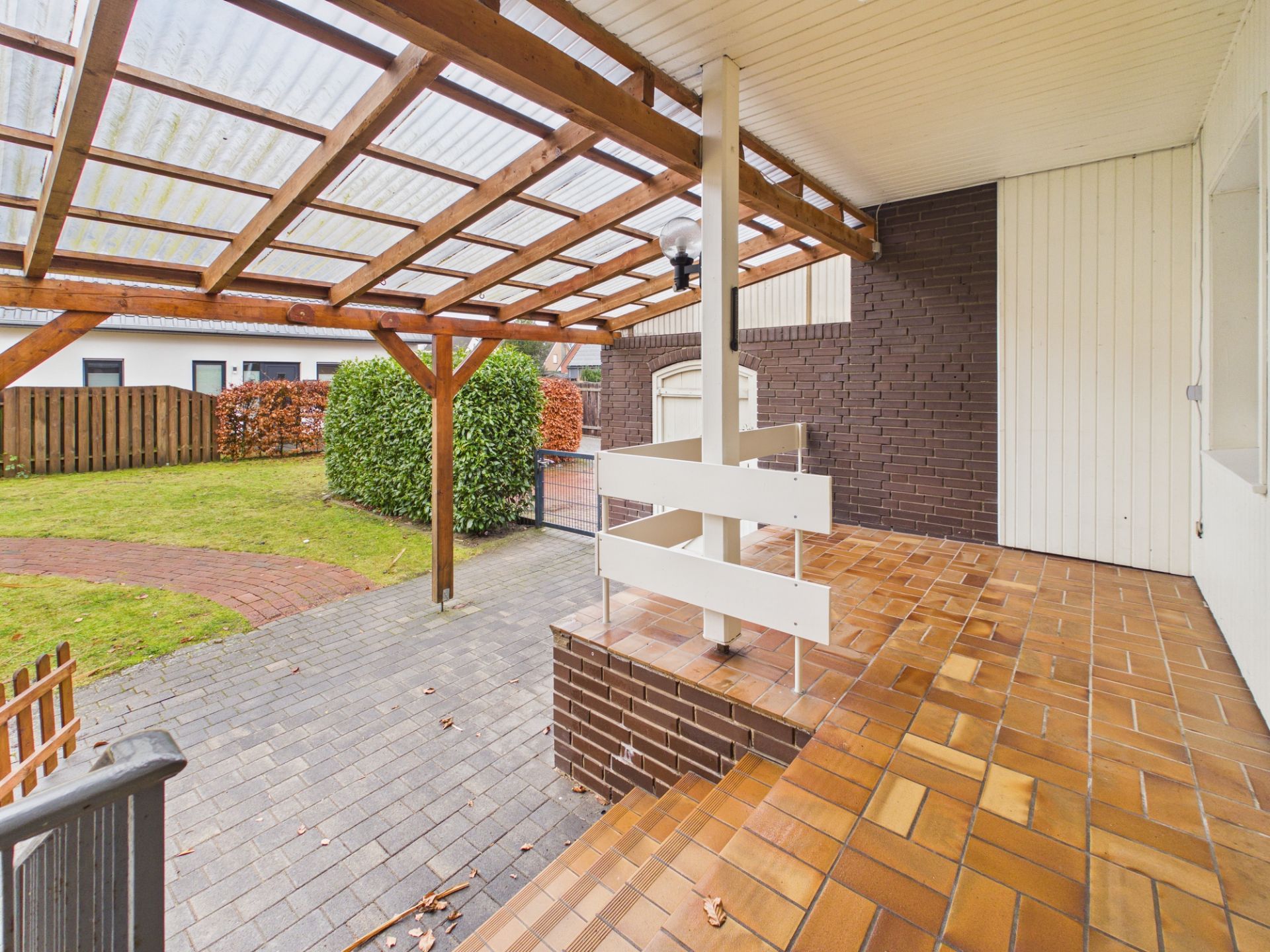 Überdachte Terrasse mit braunen Fliesen und Blick auf einen Garten mit Zaun und Hecke.