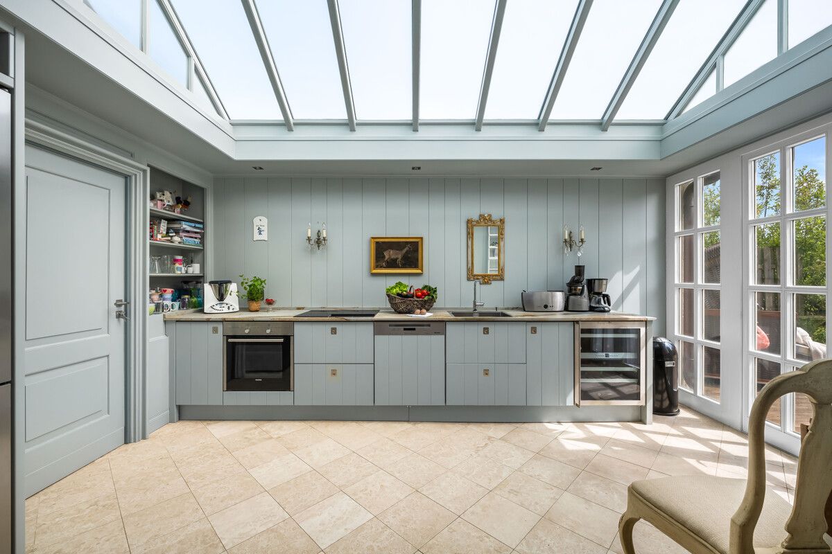 Helle Küche mit Glasdach, grauen Schränken und großen Fenstern, ausgestattet mit Backofen und Kaffeemaschine.