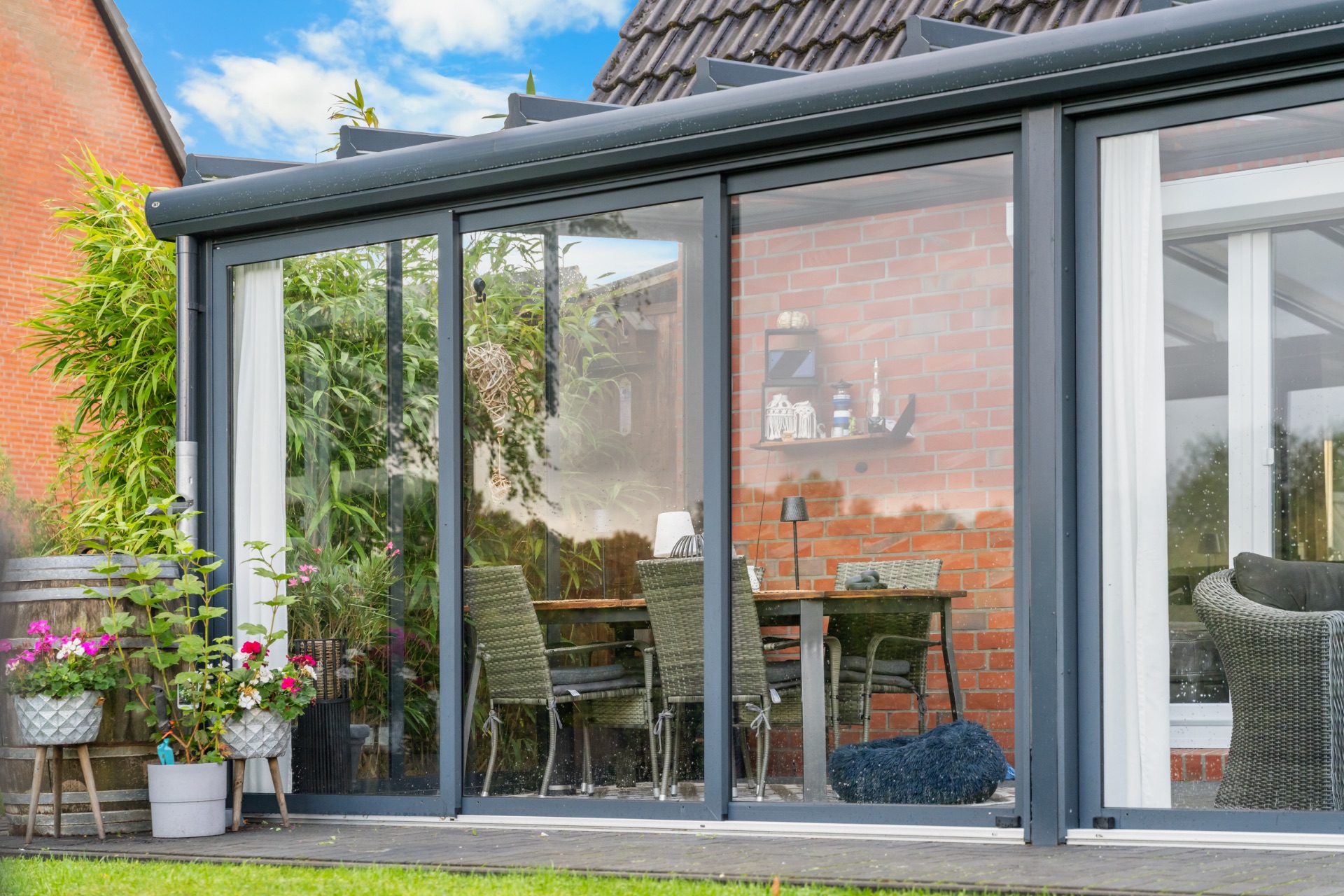 Wintergarten mit Glastüren, Rattanstühlen, Tisch und Pflanzen, Backsteinwand im Hintergrund.