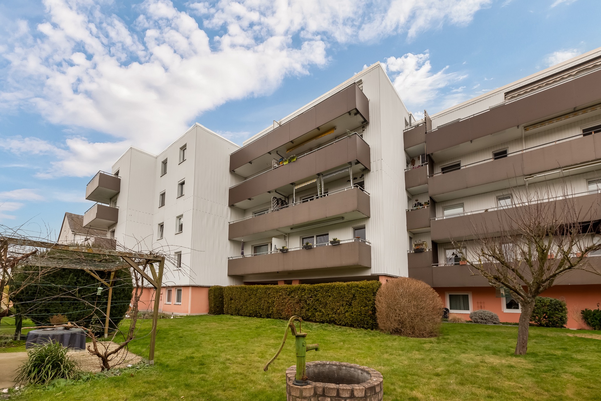 Mehrfamilienhaus mit Balkonen und Garten, im Vordergrund ein Brunnen und ein Baum.