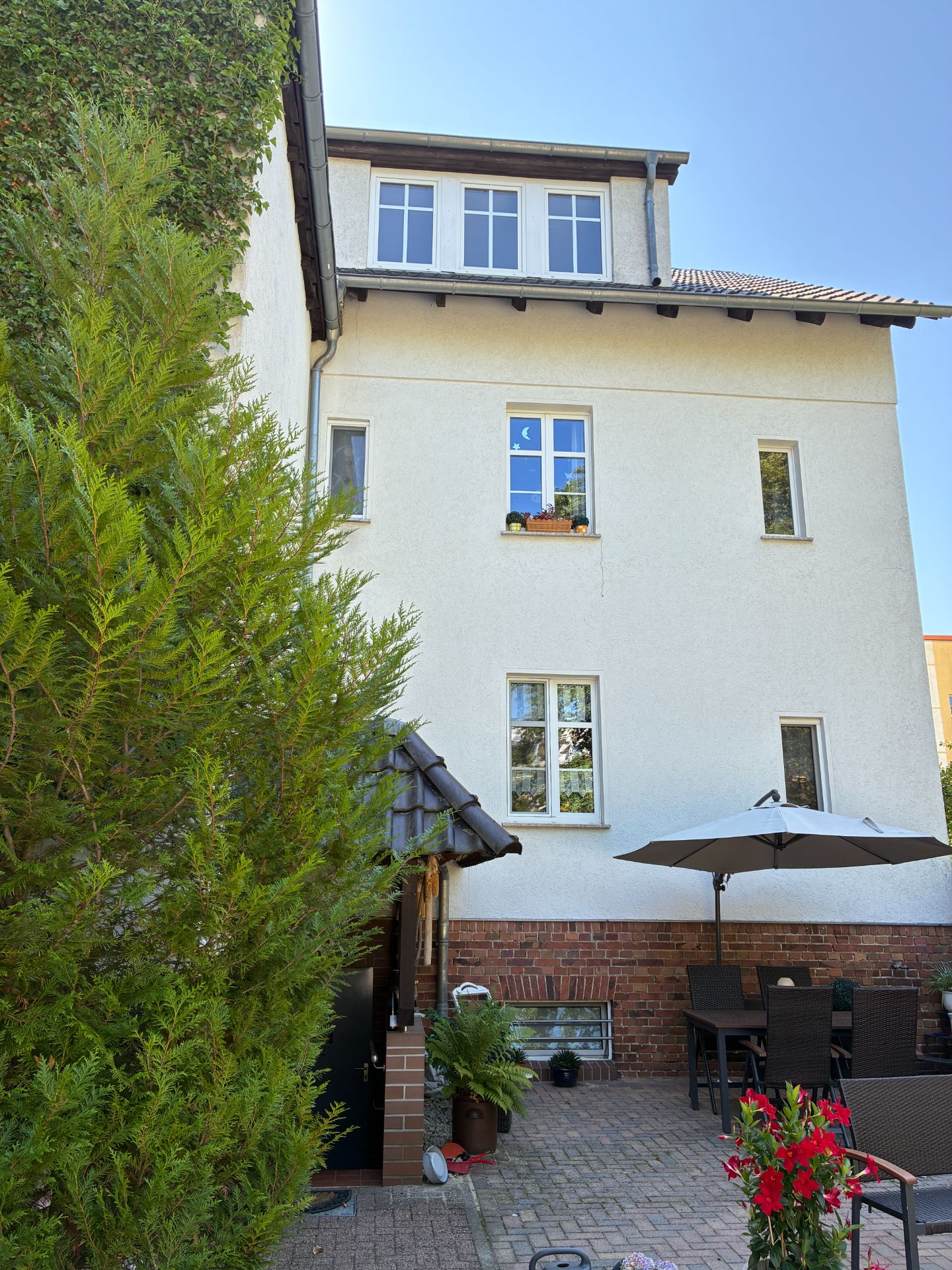 Hinterhof eines Wohnhauses mit Terrasse, Esstisch, Sonnenschirm und Pflanzen.
