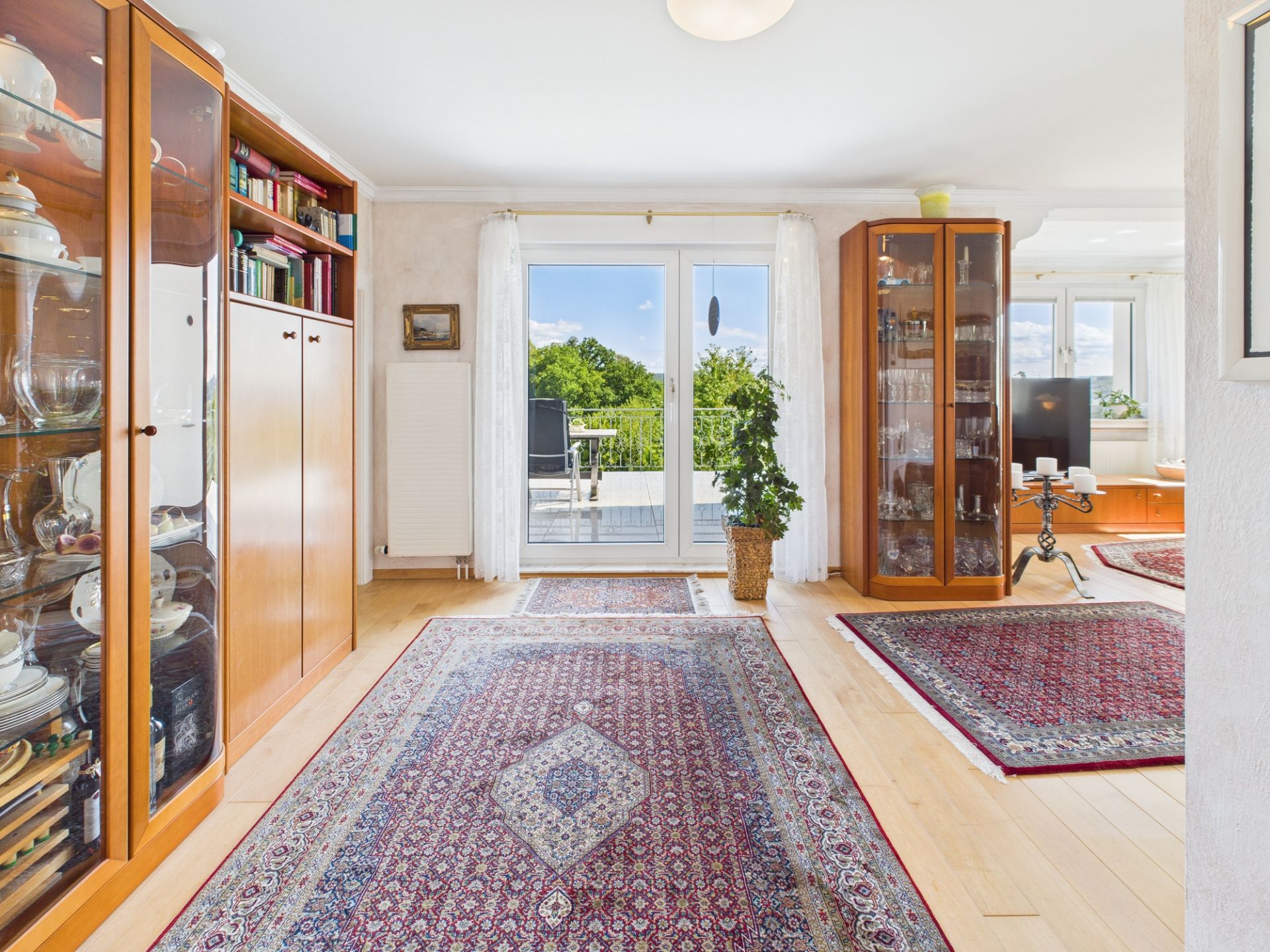 Helles Wohnzimmer mit Teppichen, Holzschränken und Zugang zu einem Balkon mit Blick ins Grüne.