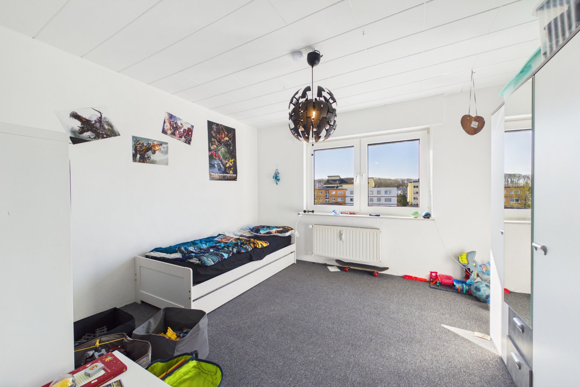Kinderzimmer mit Einzelbett, Spielzeug und Fenstern mit Blick nach draußen.