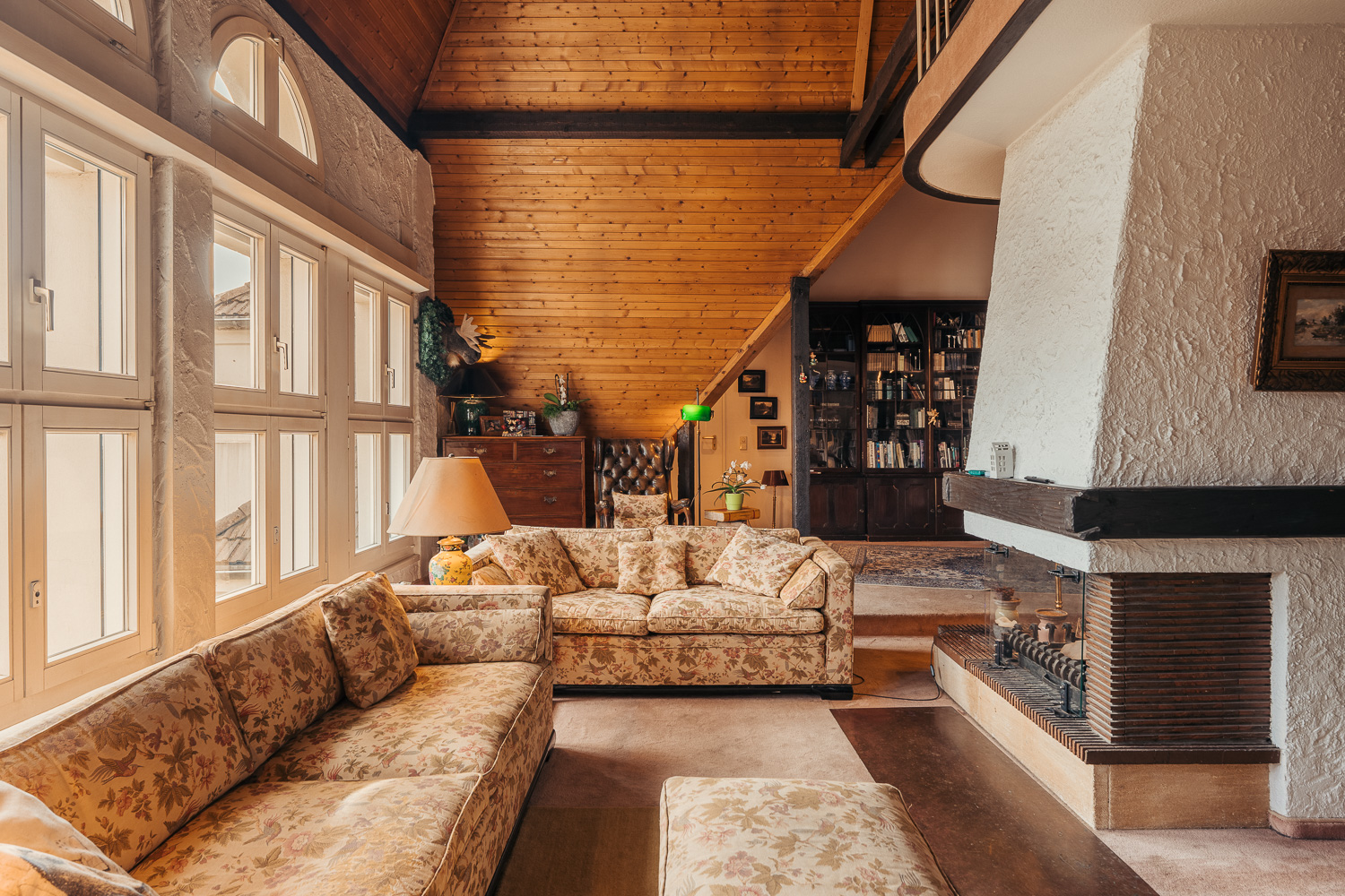 Gemütliches Wohnzimmer mit Kamin, großen Fenstern, floral gemusterten Sofas und Holzbalkendecke.