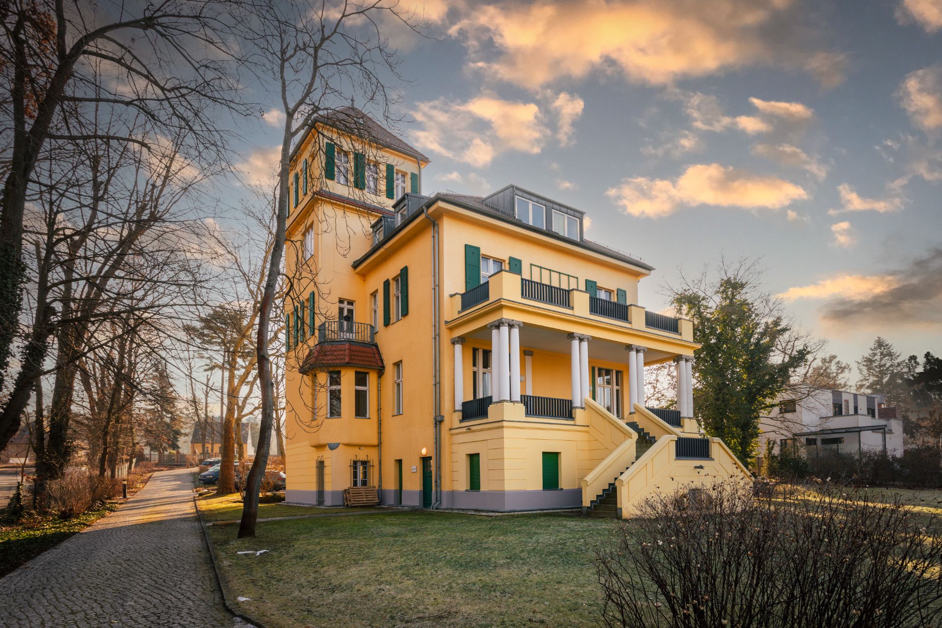Gelbes Wohnhaus mit Turm, grünen Fensterläden und Außen­treppe, umgeben von Bäumen.