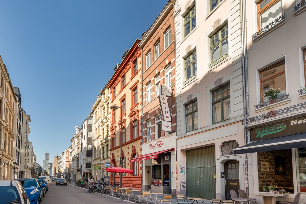 Mehrstöckige Wohngebäude an einer belebten Straße mit Straßencafés und geparkten Autos.