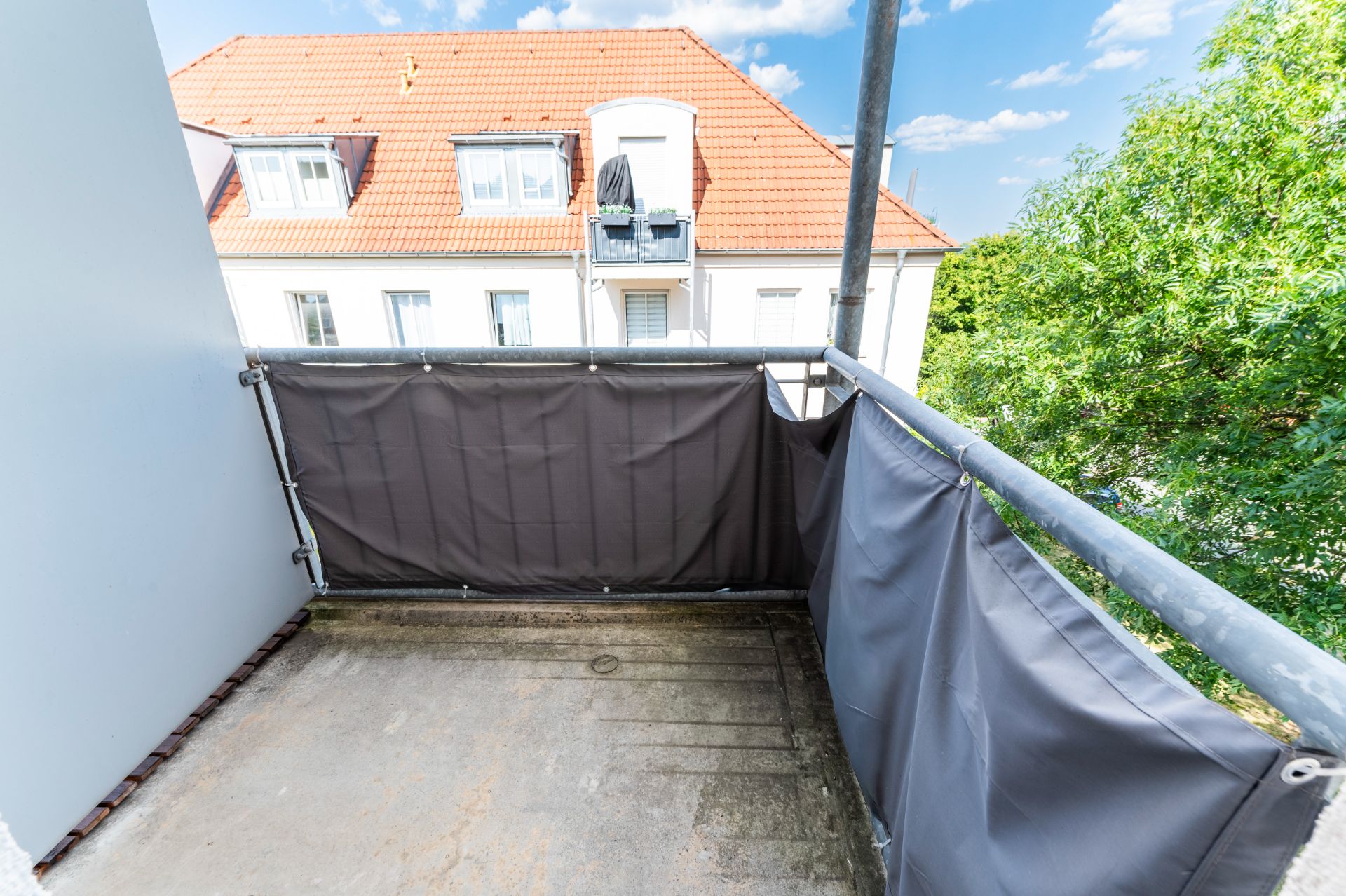 Kleiner Balkon mit grauer Verkleidung und Blick auf ein Nachbarhaus mit rotem Dach.