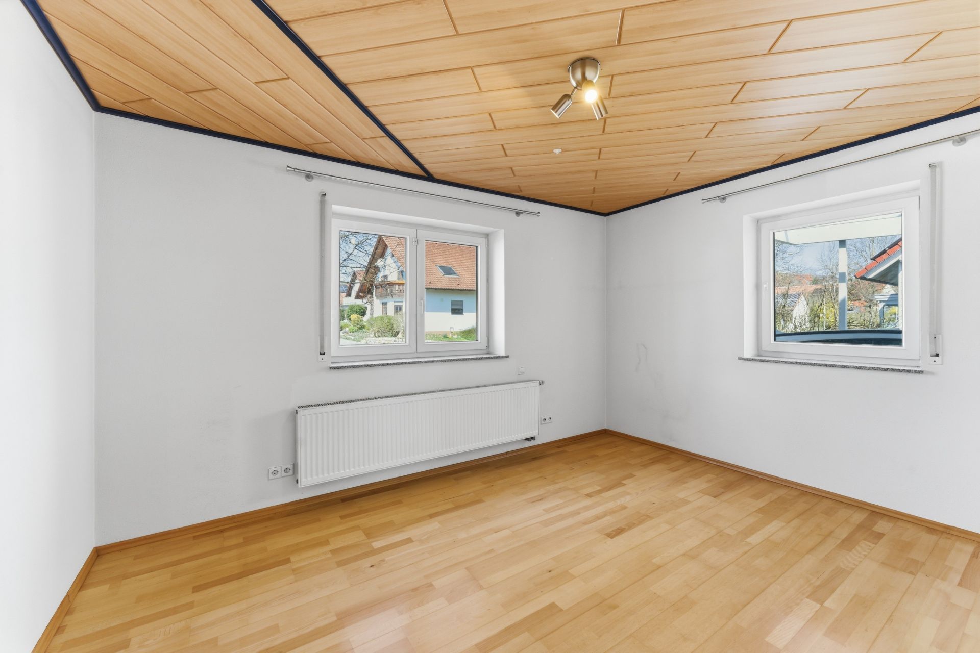 Helles Zimmer mit Holzfußboden, zwei Fenstern und Deckenleuchte.