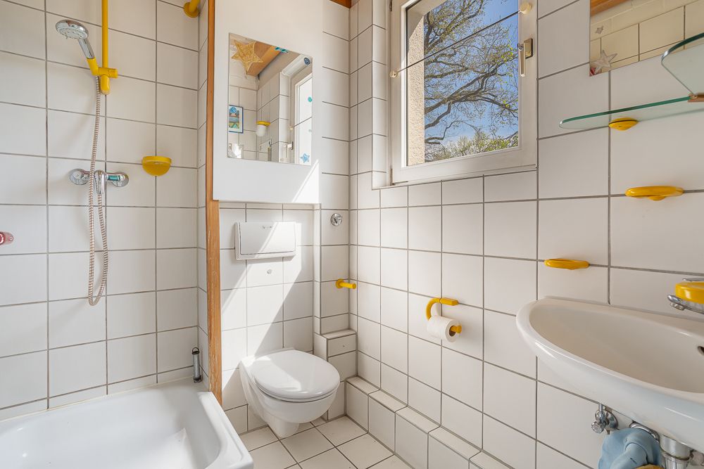 Helles Badezimmer mit Dusche, WC, Waschbecken, Spiegel und Fenster, weiße Fliesen mit gelben Akzenten.