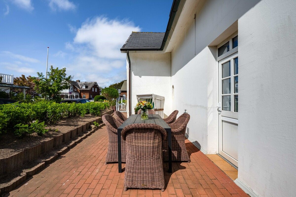 Terrasse mit Tisch, sechs Korbstühlen und Blumen neben einem weißen Haus.