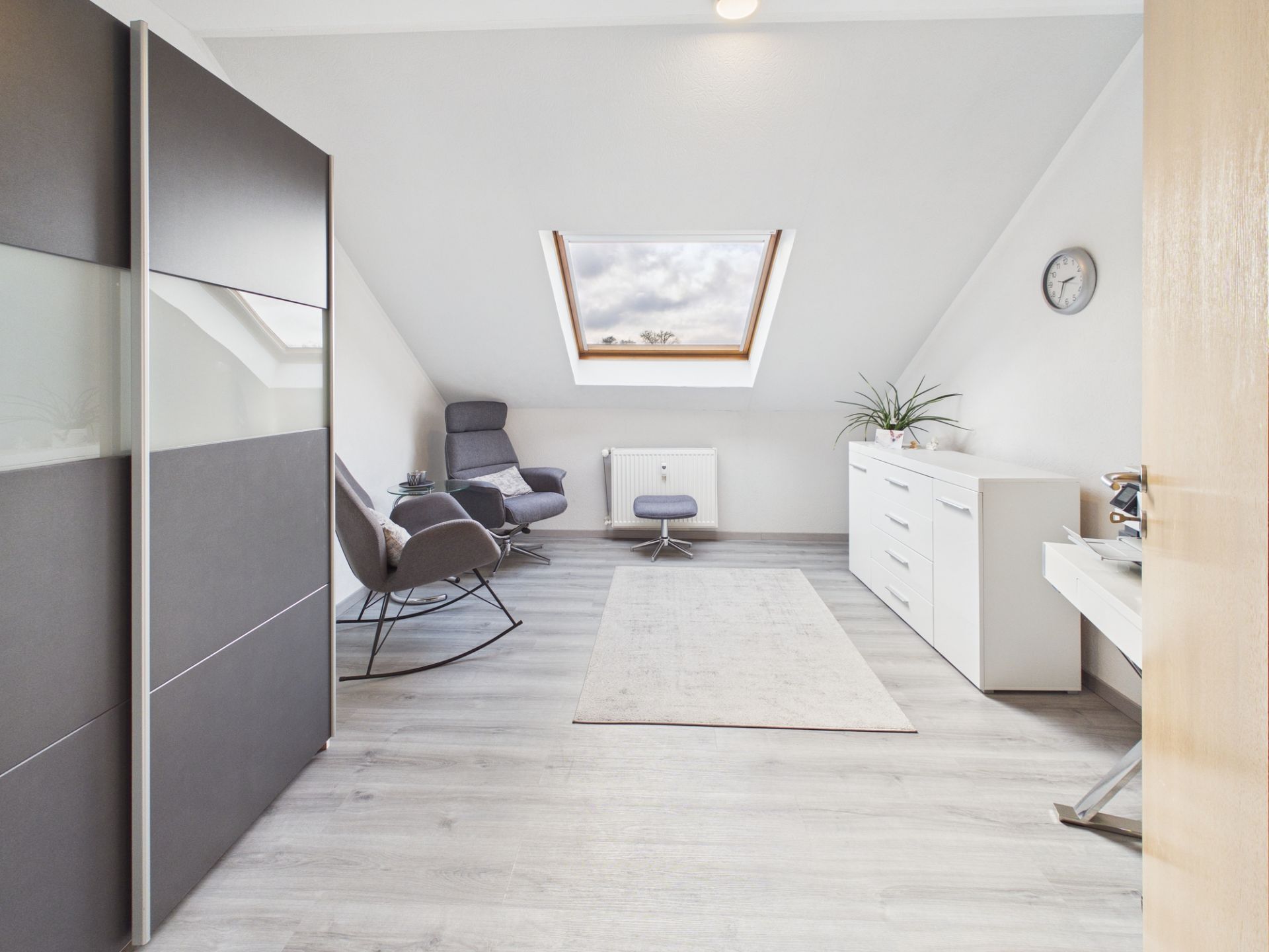 Helles Dachzimmer mit Sessel, Kommode, Schreibtisch und großem Fenster.