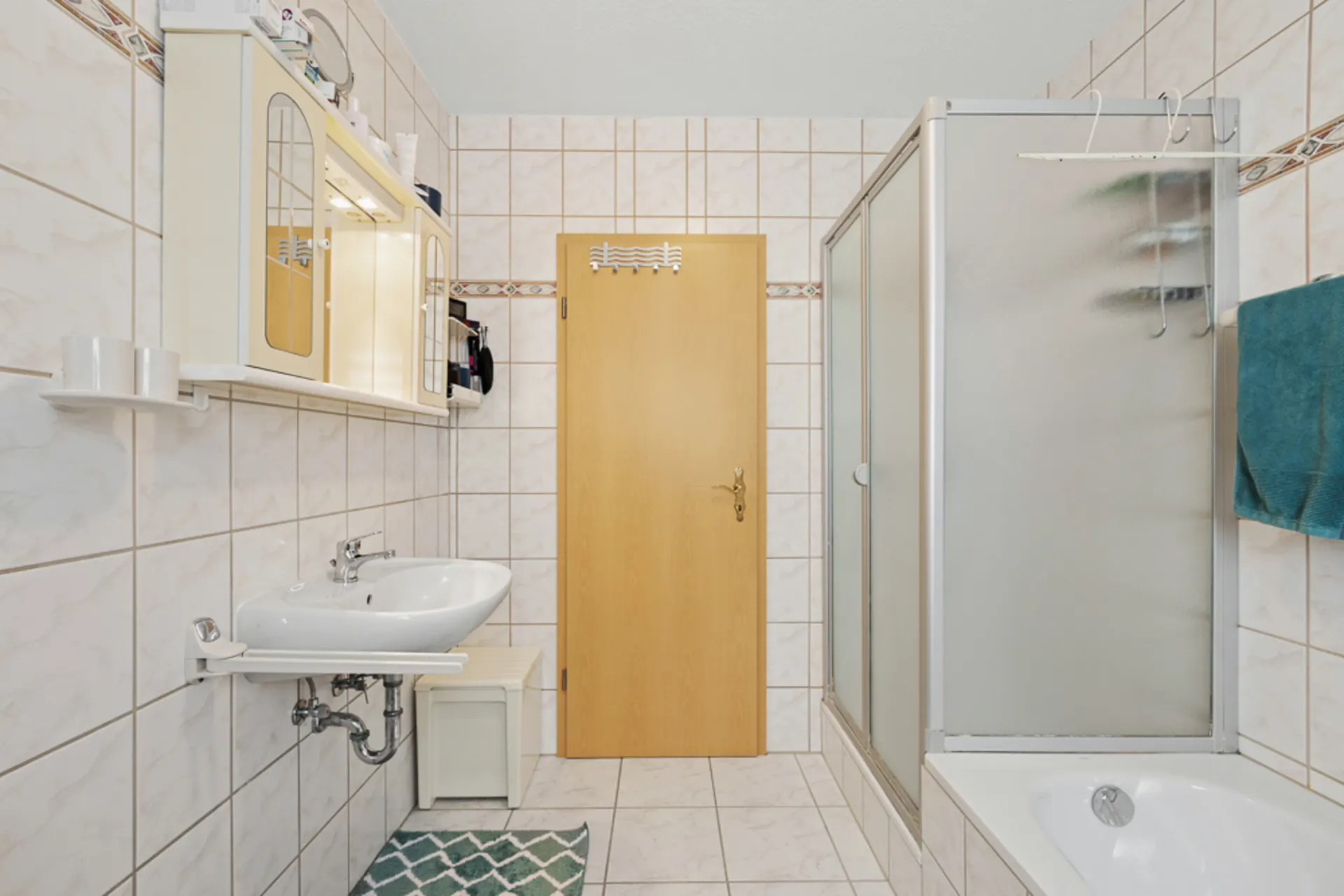 Helles Badezimmer mit Waschbecken, Spiegelschrank, Dusche, Badewanne und Holztür.