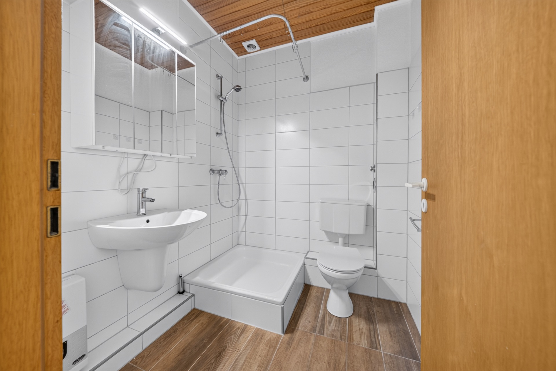 Modernes Badezimmer mit Dusche, Waschbecken, Spiegel und Toilette, weiße Fliesen und Holzboden.