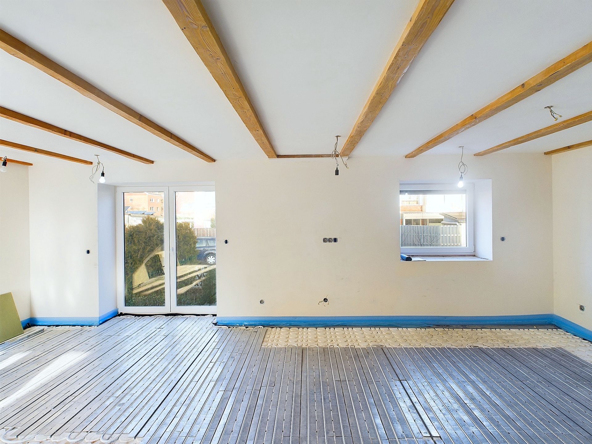 Heller Raum im Ausbau mit sichtbaren Deckenbalken und offenem Boden, Fenster und Tür nach draußen.