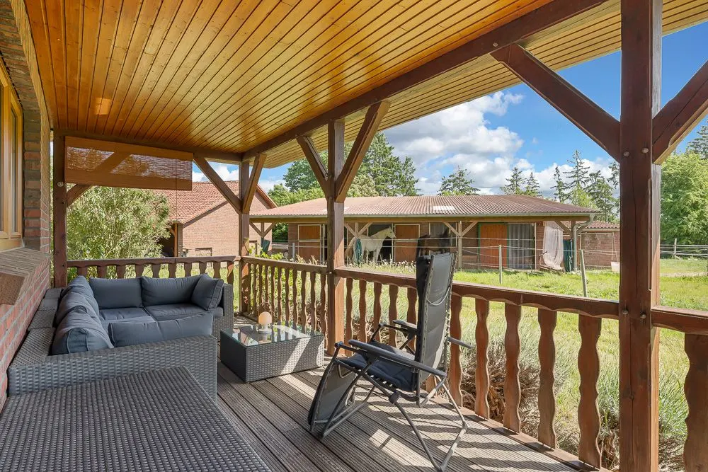 Überdachte Terrasse mit Sitzmöbeln, Blick auf Garten und Stall mit Pferden.