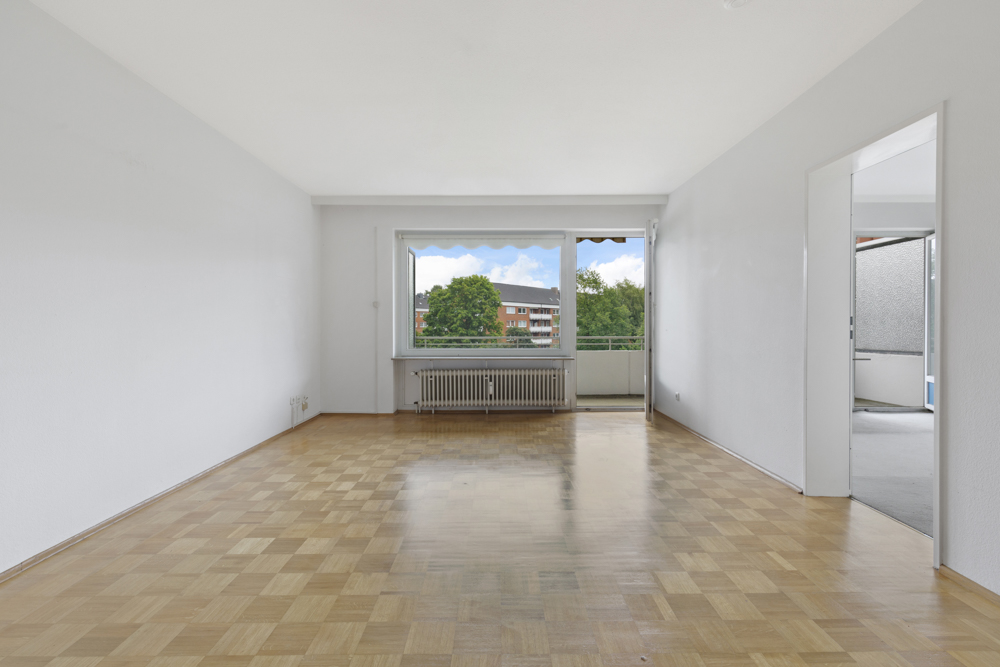 Leeres Wohnzimmer mit Parkettboden, großem Fenster und Zugang zu einem Balkon.