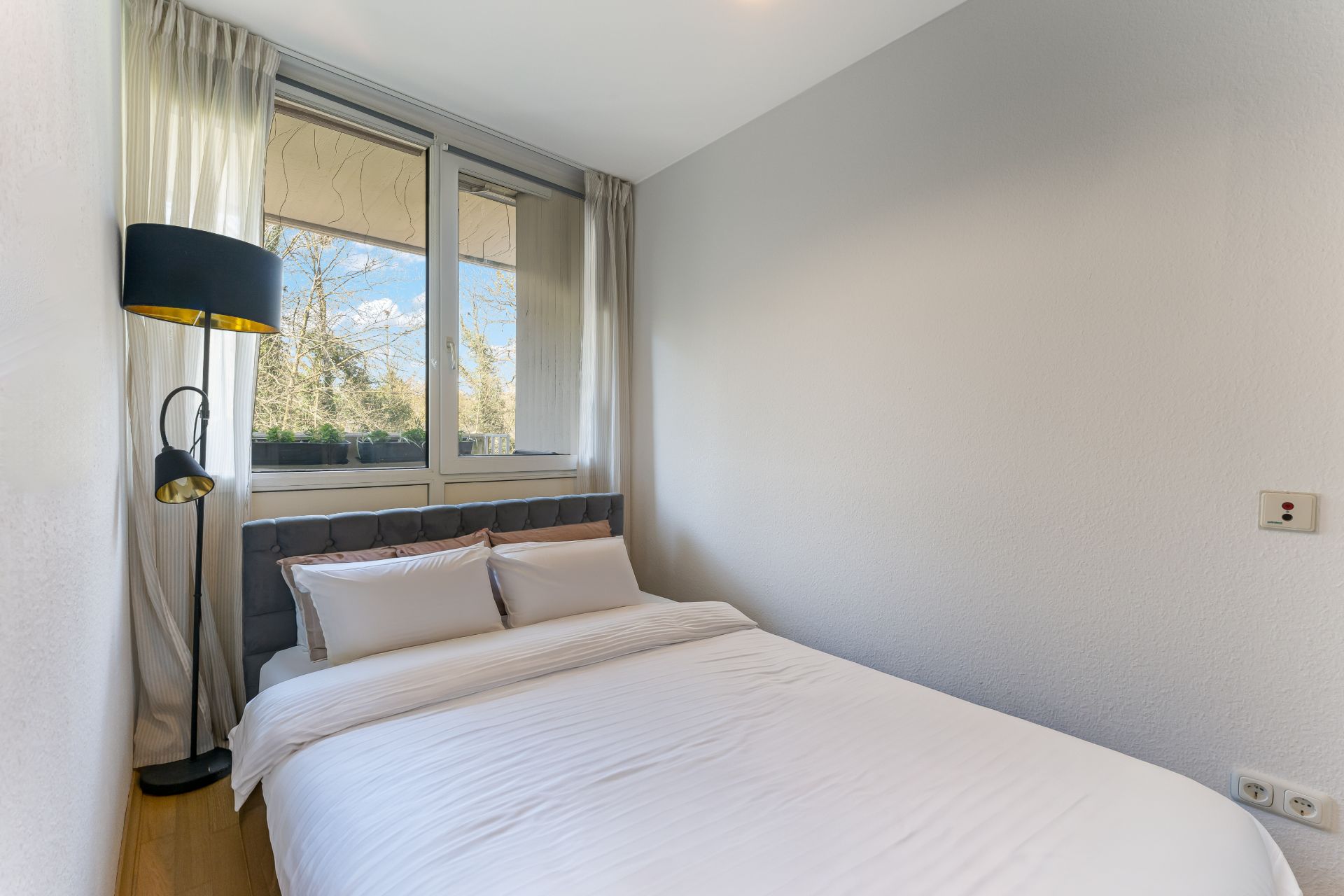 Kleines Schlafzimmer mit Doppelbett, Fenster, weißen Vorhängen und Stehlampe.