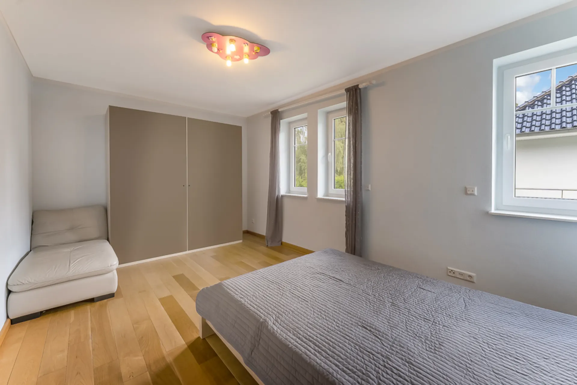 Helles Schlafzimmer mit großem Fenster, grauem Bett, Schrank und Sessel auf Holzfußboden.