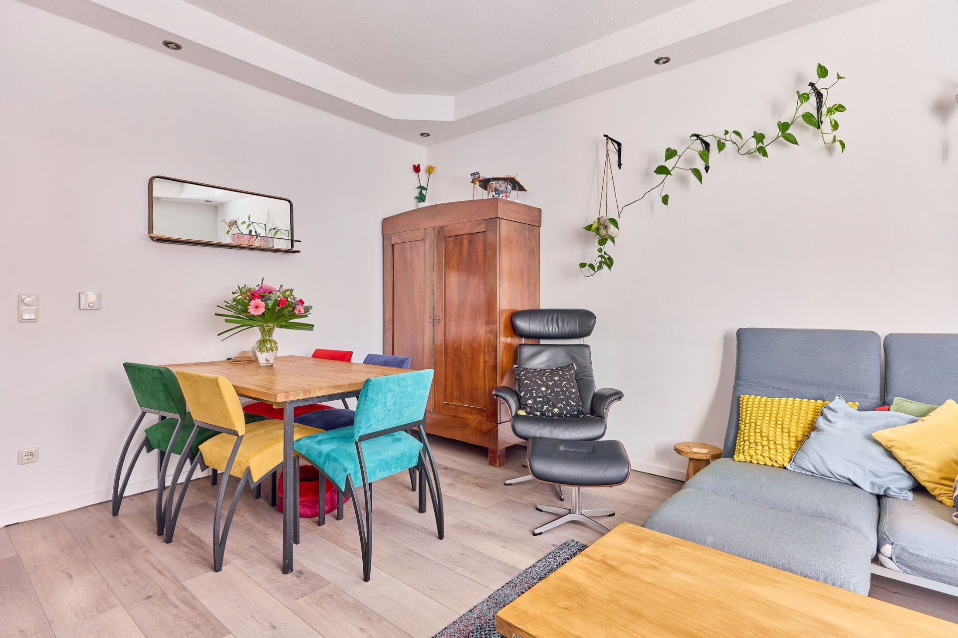 Wohnzimmer mit bunten Stühlen, Holzschrank, Sessel, Sofa und hängenden Pflanzen an der Wand.