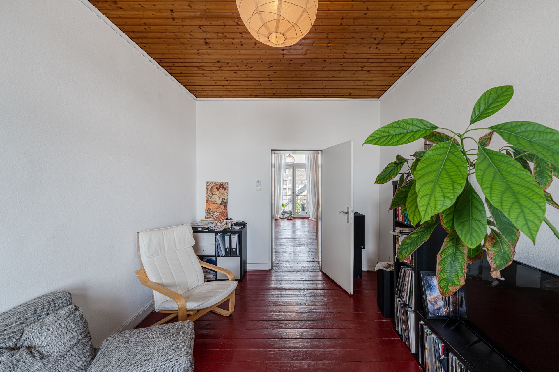 Wohnzimmer mit rotem Holzboden, weißem Sessel, Regal, Pflanze und Blick in ein weiteres Zimmer.