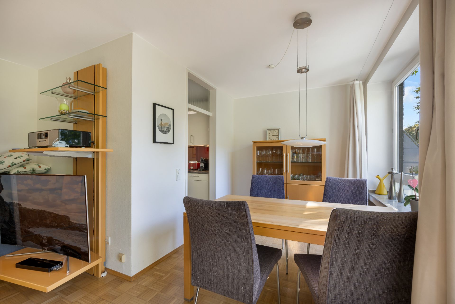 Essbereich mit Holztisch und vier grauen Stühlen, großer Fensterfront und Schrank mit Glastüren.