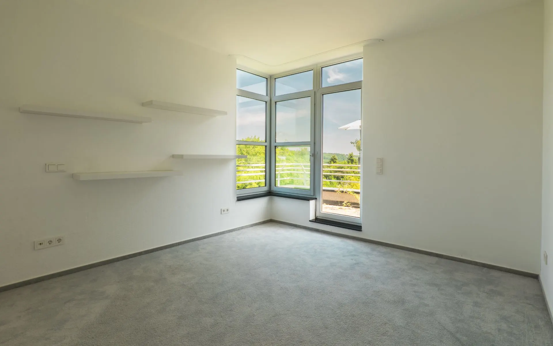 Leeres Zimmer mit Teppichboden, mehreren Wandregalen und großen Fenstern mit Blick nach draußen.