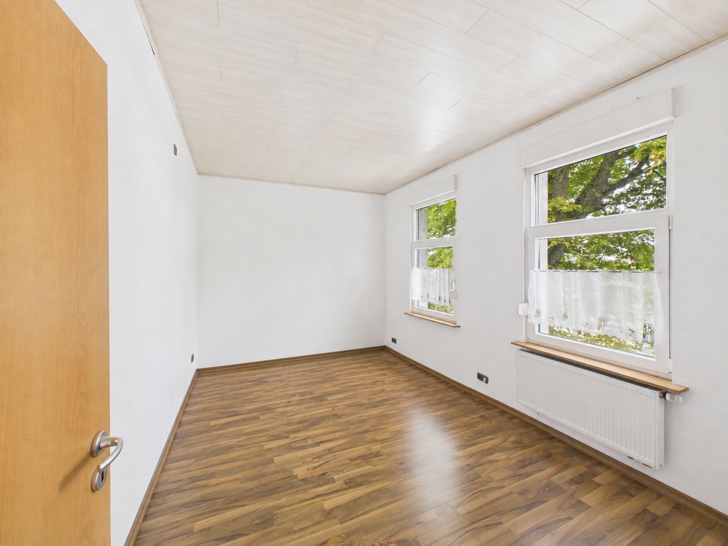 Leerer Raum mit Holzfußboden, weißen Wänden und zwei Fenstern mit Blick ins Grüne.