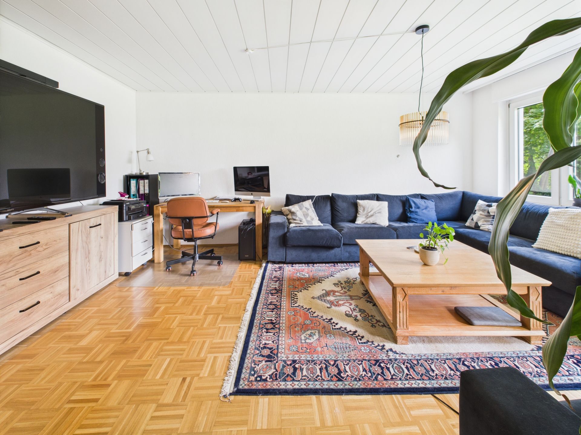Helles Wohnzimmer mit großem Ecksofa, Holztisch, Teppich, Fernseher und Schreibtisch am Fenster.