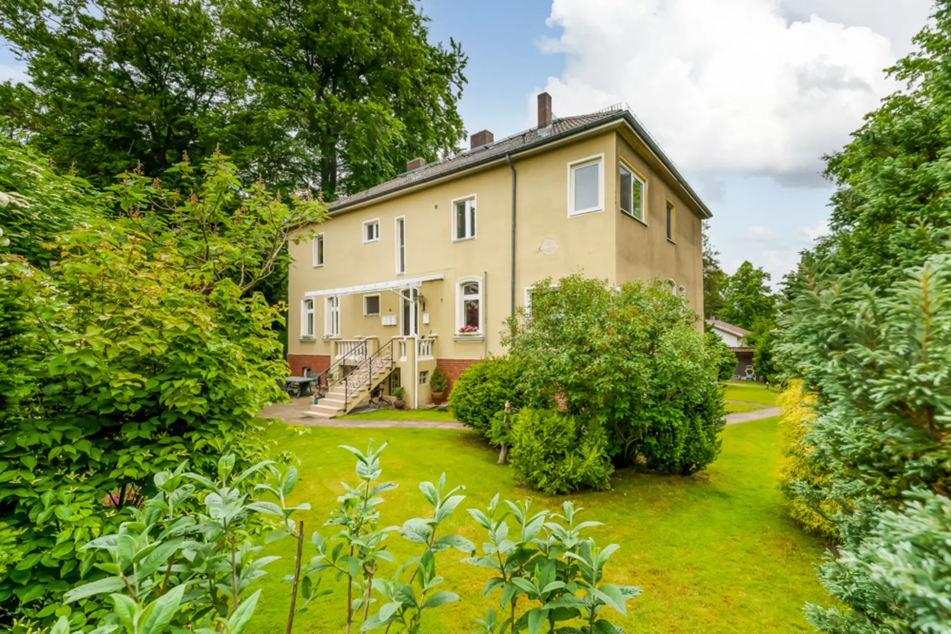 Zweistöckiges Wohnhaus mit hellem Putz, umgeben von grünem Garten und Bäumen.