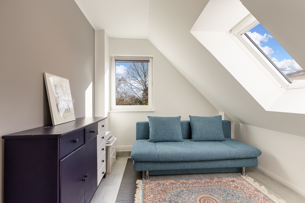 Kleines Zimmer mit blauem Sofa, Kommode, Teppich und zwei Fenstern unter Dachschräge.