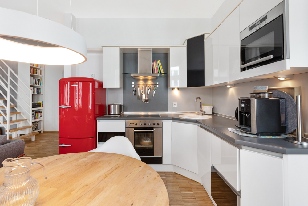 Moderne Küche mit rotem Kühlschrank, Backofen, Kaffeemaschine und rundem Holztisch.