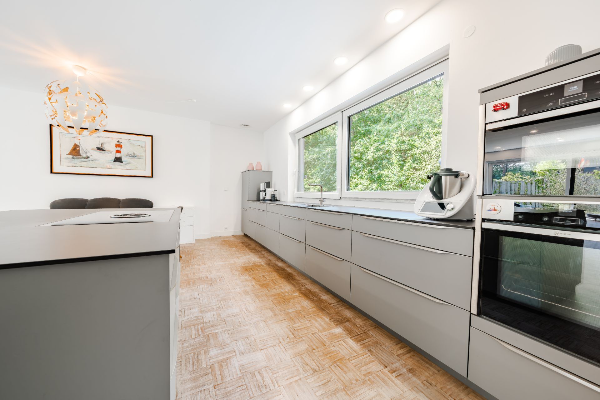 Helle, moderne Küche mit grauen Schränken, großem Fenster und Einbaugeräten.