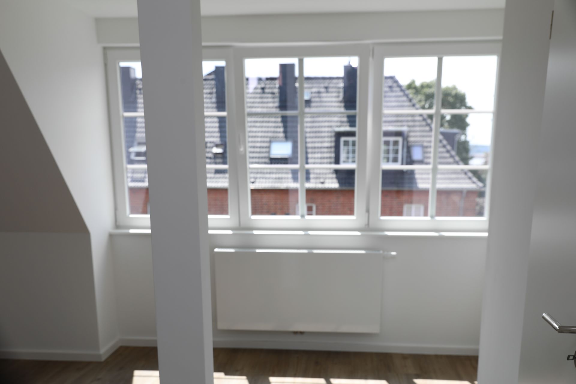 Helles Zimmer mit großen Fenstern, Blick auf Nachbarhäuser und Heizkörper unter dem Fenster.