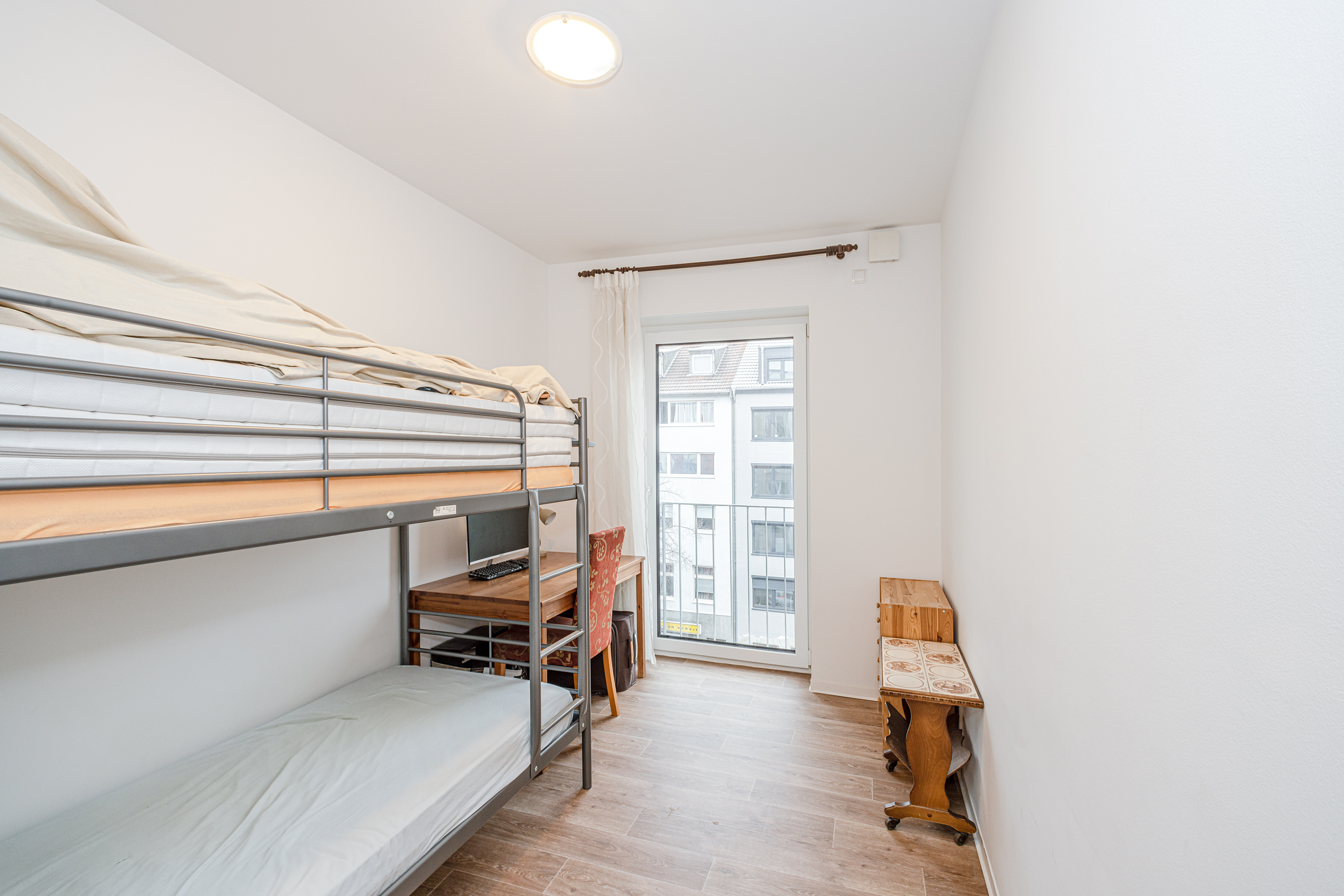 Helles Zimmer mit Etagenbett, Schreibtisch, Stuhl und Fenster mit Blick auf andere Häuser.