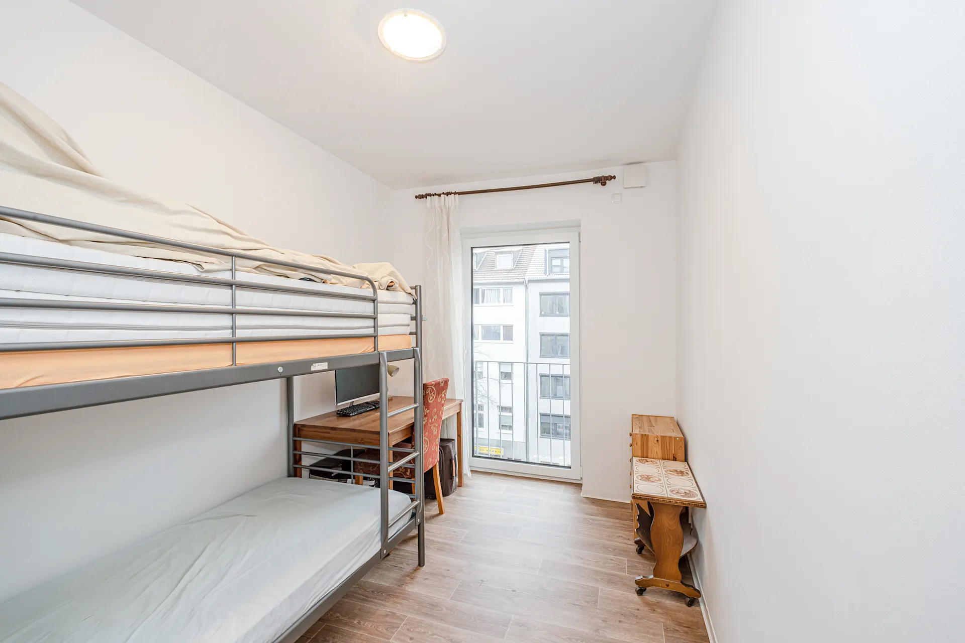 Helles Zimmer mit Etagenbett, Schreibtisch, Stuhl und Fenster mit Blick auf andere Häuser.