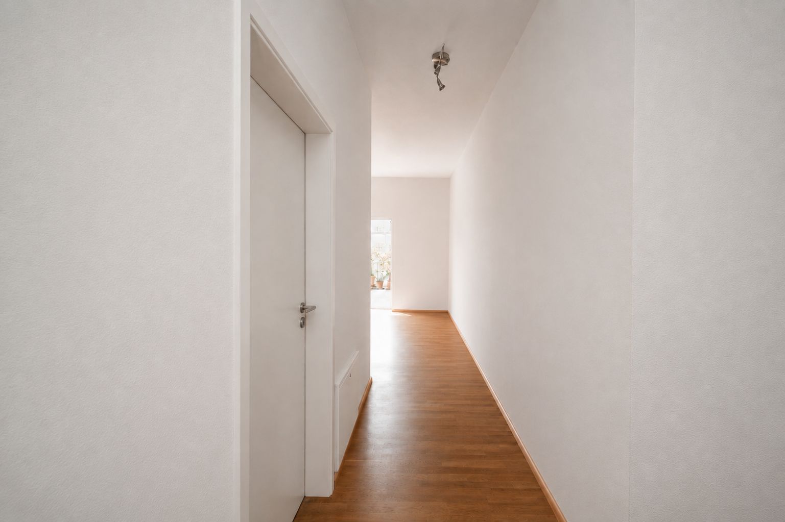 Heller Flur mit weißer Tür und Holzfußboden, am Ende ein Fenster mit Tageslicht.