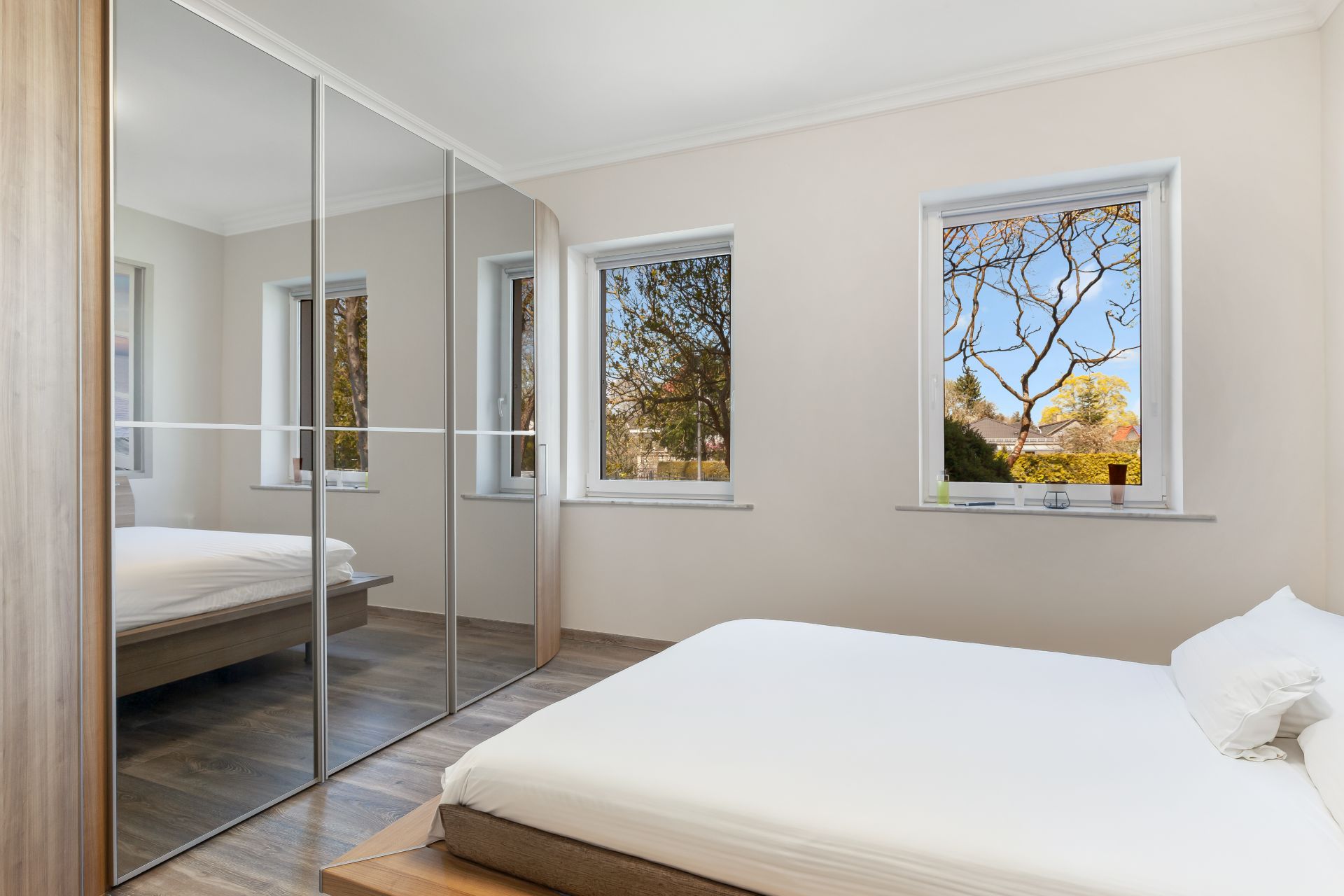 Helles Schlafzimmer mit Doppelbett, drei Fenstern und großem Kleiderschrank mit Spiegeln.