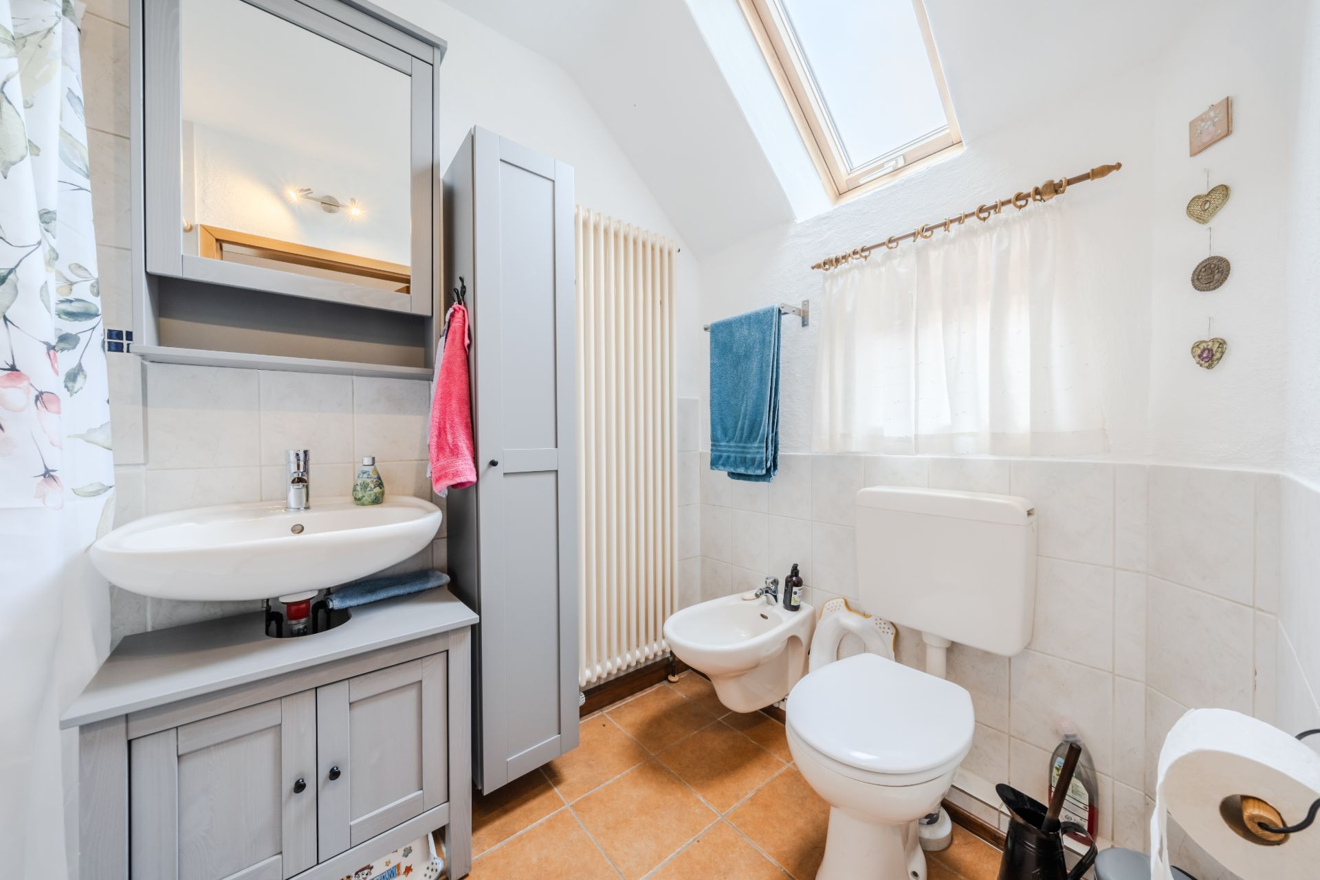 Helles Badezimmer mit Waschbecken, Spiegel, Toilette, Bidet, Fenster und grauem Schrank.
