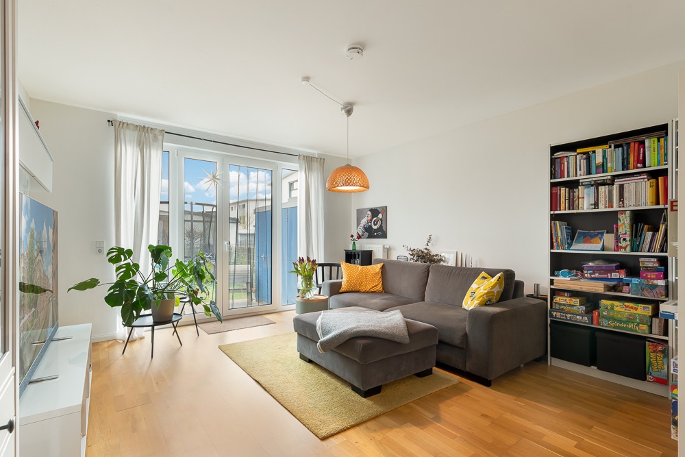 Helles Wohnzimmer mit Ecksofa, Bücherregal, Pflanzen und großem Fenster zum Garten.