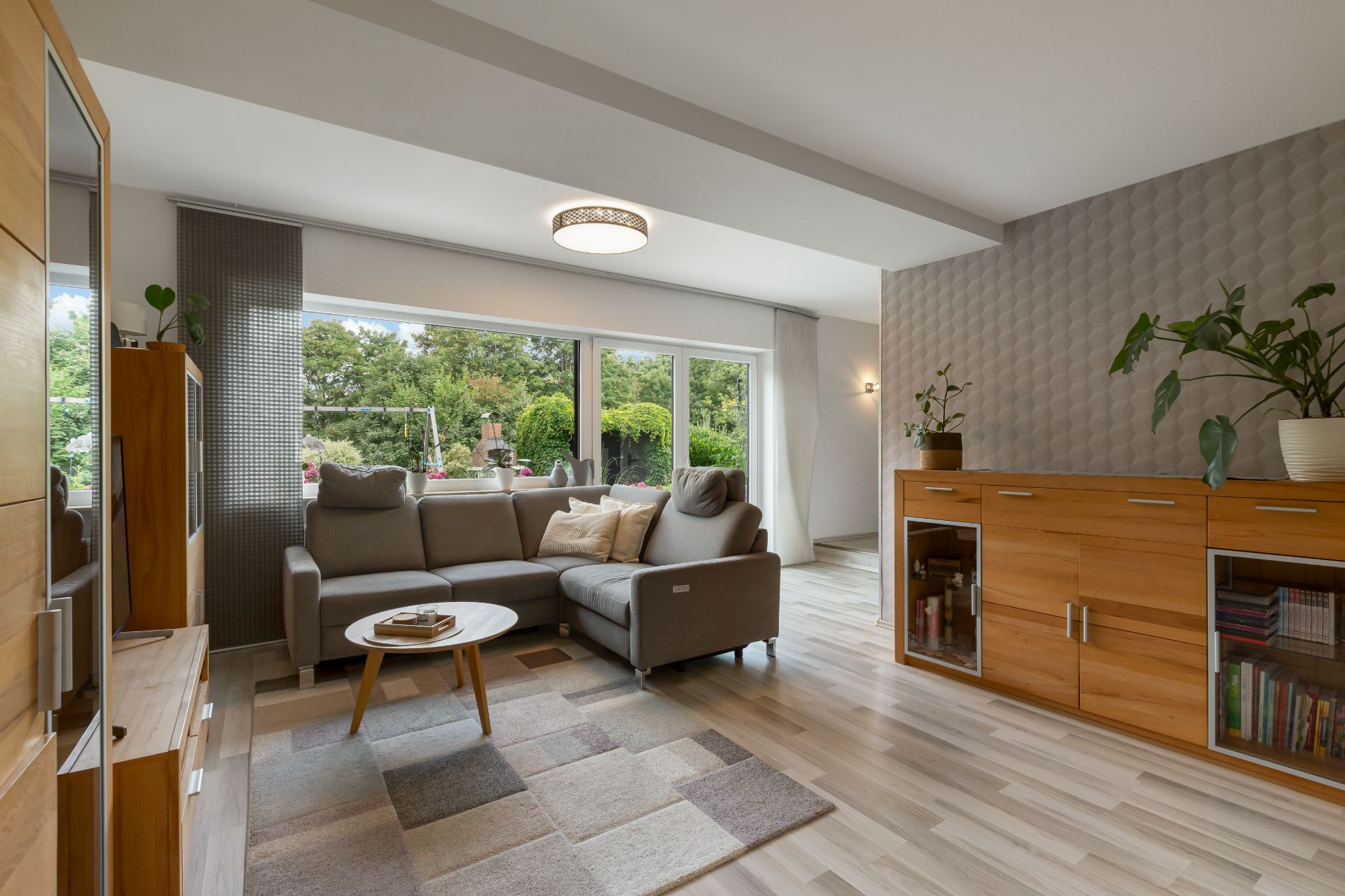 Modernes Wohnzimmer mit grauem Sofa, Holzschrank, Teppich und großen Fenstern zum Garten.