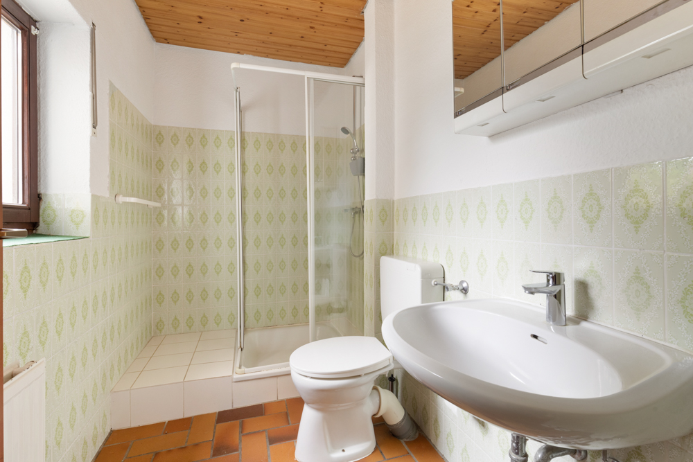 Helles Bad mit Dusche, Toilette und Waschbecken, grüne Fliesen, Holzdecke.