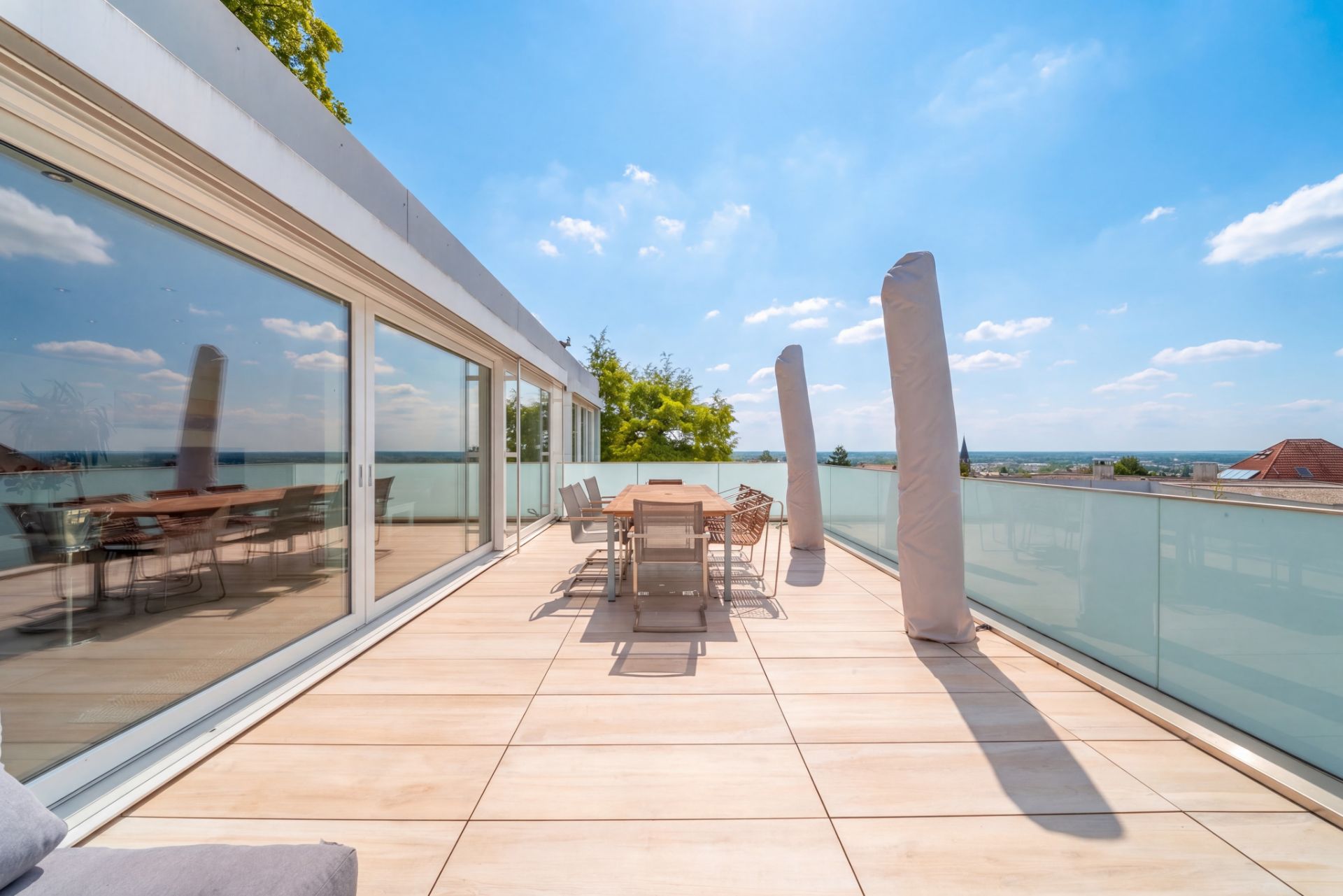 Große Terrasse mit Esstisch, Stühlen und Glasgeländer, daneben große Fensterfront.
