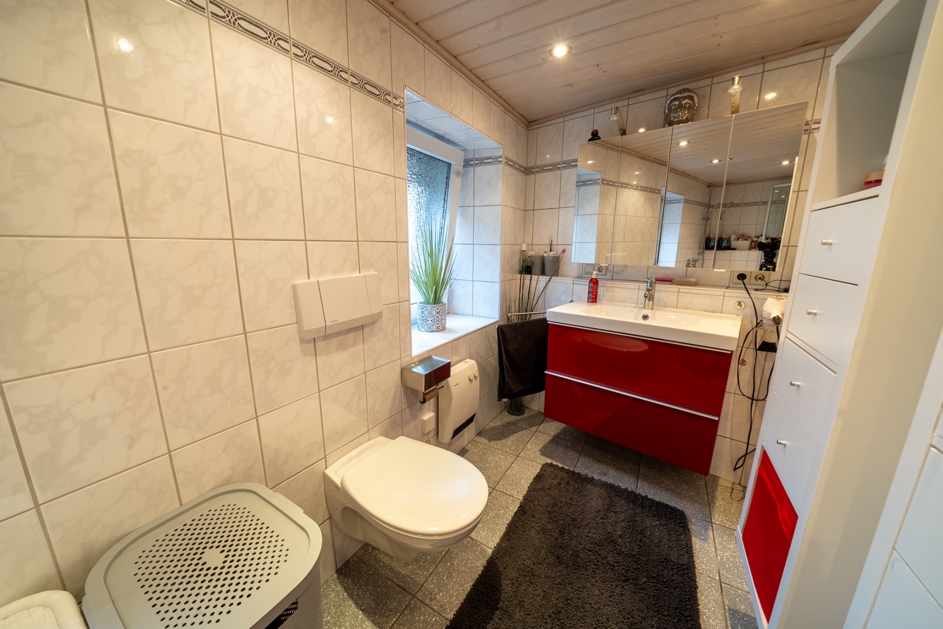 Modernes Badezimmer mit weißem WC, rotem Waschtisch, großem Spiegel und grauem Teppich.
