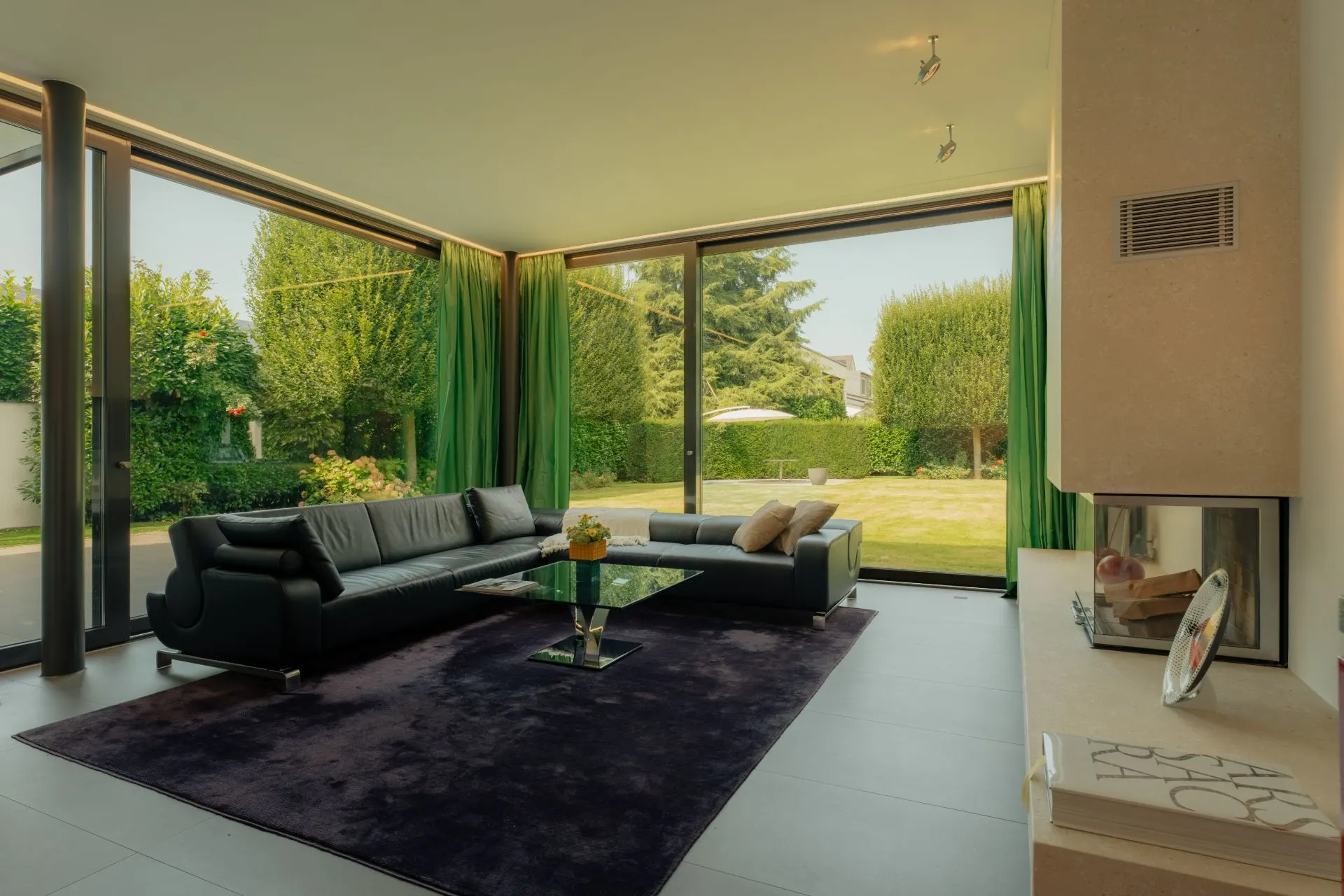 Modernes Wohnzimmer mit schwarzem Sofa, Glastisch, Kamin und großen Fenstern zum Garten.
