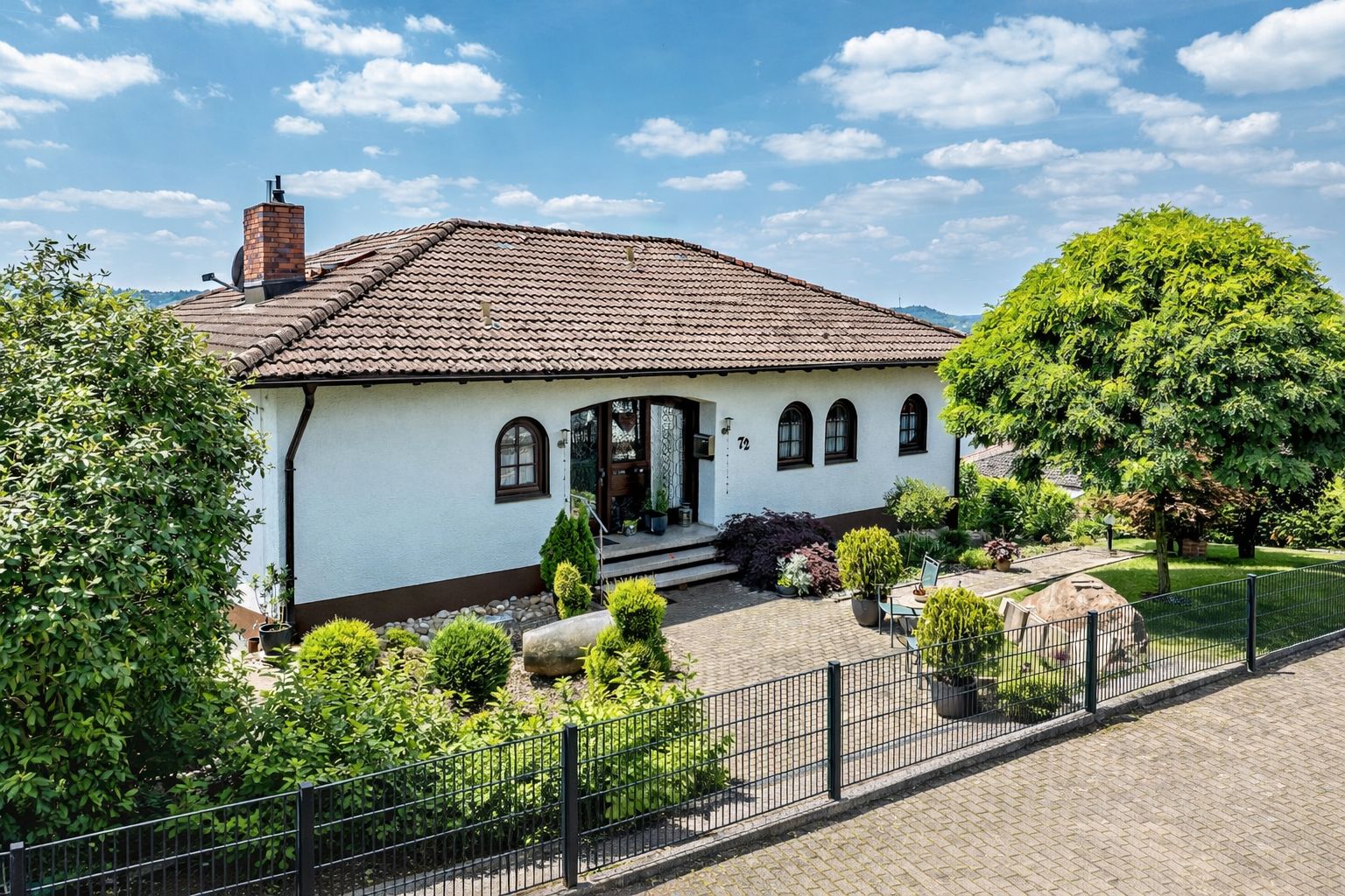 Einfamilienhaus mit braunem Dach, Vorgarten und Zaun an einer gepflasterten Straße.