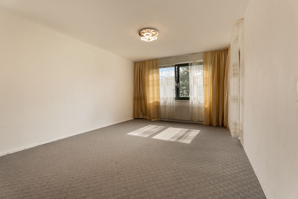 Leeres Zimmer mit Teppichboden, Fenster und gelben Vorhängen, beleuchtet von einer Deckenlampe.